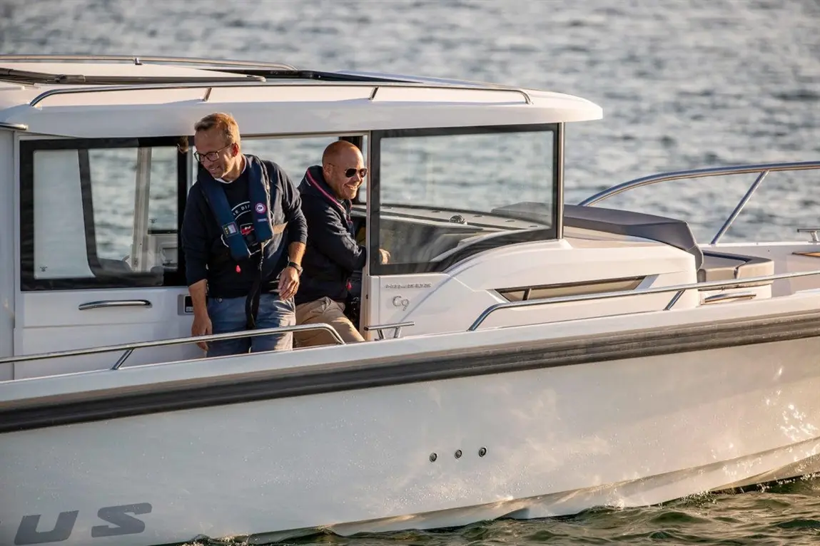 Two men on a modern boat, one standing and laughing while the other sits at the helm, with water in the background.